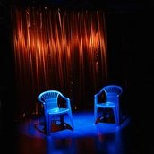 Zwei leere blaue Plastikstühle stehen im Bühnenlicht vor einem gold schimmernden Vorhang. Der restliche Raum liegt im Dunkeln.