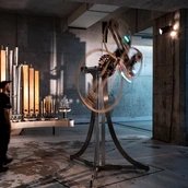 In einem kargen Raum stehen zwei Objekte. Eines besteht aus unterschiedlichen, nebeneinander aufgereihten Orgelpfeifen, das andere aus einem Stativ und sich drehenden Kreisen.