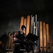 Eine Person mit hochgesteckten lockigen Haaren bedient ein Mischpult. Er steht vor einer Installation aus Orgelpfeifen.