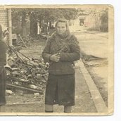 Das Foto zeigt zwei junge Mädchen vor dem Hintergrund von Trümmern. Das Mädchen links trägt ein dunkles Kleid und ein Kopftuch. Das Mädchen rechts trägt ein dunkleres Kleid, Strümpfe und einen gehäkelten Schal. Der Hintergrund deutet auf Zerstörung u
