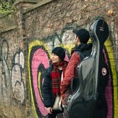 Zwei Personen stehen vor einer Backsteinwand, die mit Graffiti besprayt ist. Bei der linken Person handelt es sich um die Gastgeberin Antonia Regis. Die rechte Person ist nur von hinten zu sehen, sie trägt ein Cello-Koffer.