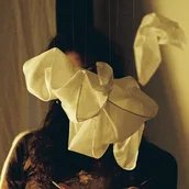 Das Bild zeigt eine Frau, deren Gesicht teilweise von einer hängenden Installation aus weißen Stoffelementen verdeckt wird, was eine mysteriöse und intime Atmosphäre erzeugt.