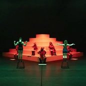 Performer:innen auf Hockern und Musiker:innen in roten Kapuzen posieren vor einer stufenförmigen weißen Plattform im grünen und roten Licht.