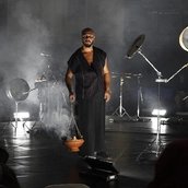 Ein Mann in einem ärmellosen schwarzen Outfit steht auf einer dunklen, rauchigen Bühne und hält einen Ton-Räuchergefäß in der Hand, aus dem Rauch aufsteigt. Um ihn herum stehen unter sanftem weißem Licht Percussion-Instrumente und Mikrofone, während 