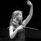 Dirigentin Mirga Gražinytė-Tyla hält in der rechten Hand den Taktstock und hebt den linken Arm vor dem Orchester hoch.