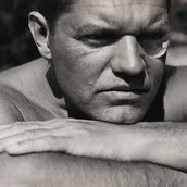 Schwarz-Weiß-Fotografie von Raoul Hausmann, der mit runder Sehhilfe im linken Auge, den Kopf auf den verschränkten Armen aufgelegt, an den Betrachtenden vorbei in die Ferne blickt.