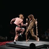 Zwei Personen in erdfarbenen Anzügen stampfen. Sie haben Stöcke mit Glocken in der Hand.