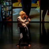 Eine barfüßige Performer:in mit blondem Haar tanzt allein auf dunkler Bühne, im Hintergrund sind großformatige Videoausschnitte mit Innenraumszenen zu sehen.