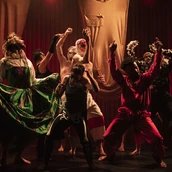 Eine Gruppe von Darstellern in farbenfrohen, dramatischen Kostümen posiert dynamisch auf einer schwach beleuchteten Bühne mit roten Vorhängen und drückt unter der Theaterbeleuchtung intensive Emotionen und Bewegungen aus.