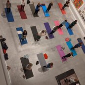 Foto: Einige Personen stehen auf bunten Matten und machen Yoga in einem Ausstellungsraum.