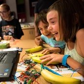 Kinder sitzen an einem Tisch vor einem Laptop. Auf dem Bildschirm ist ein Spiel mit einem Rennauto zu sehen. Die Kinder steuern das Spiel mit Bananen, die auf dem Tisch liegen und über Kabel und ein Gerät mit dem Laptop verbunden sind.