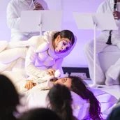 Zwei weiß gekleidete und geschminkte Frauen spielen etwas auf einer Bühne im Hintergrund spielen Musiker