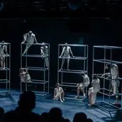 Auf einer dunklen Bühne stehen vier silberne Gerüste nebeneinander, auf denen sich mehrere Tänzer*innen stehend oder sitztend positionieren. Sie tragen weiße, schlichte Kostüme.