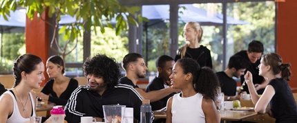 Un grupo de personas está sentado en las mesas de una cafetería muy iluminada del hotel Citylight. Charlan y disfrutan de la comida. Una mujer observa desde el fondo. Los grandes ventanales proporcionan luz natural.
