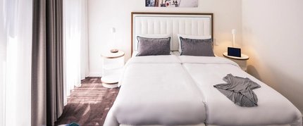 Ein modernes Hotelzimmer mit einem Doppelbett, zwei Nachttischen und einem teilweise geöffneten, deckenhohen Fenster mit Vorhängen. An der Wand über dem Bett ist ein Foto von einem Boot angebracht, und auf dem Bett liegt eine Decke.