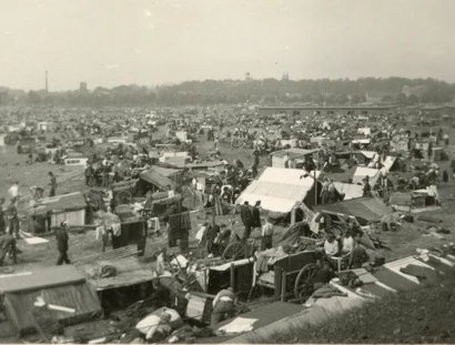 Das Schwarz-Weiß-Foto zeigt eine große Ansammlung von Zelten und Wagen auf einer weitläufigen Fläche. Viele Menschen sind zu sehen, die sich in und um die Behausungen herum bewegen. Im Hintergrund ist eine Bebauung mit Bäumen und Gebäuden erkennbar. 