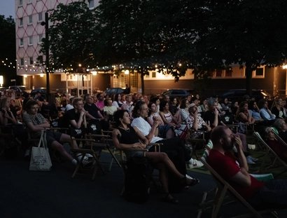 Viele Menschen sitzen in Stuhlreihen auf dem Vorplatz der Berlinischen Galerie und blicken nach vorne auf eine Leinwand, die nicht auf dem Bild zu sehen ist. Es dämmert langsam.