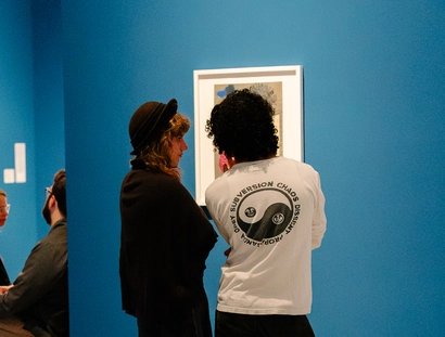 Rückenansicht von zwei Personen, die vor einer Collage von Raoul Hausmann stehen, die an einer leuchtend blauen Wand hängt.