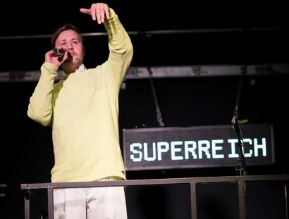 Eine Person in einem gelben Pulli und mit einer weissen Hose redet in ein Mikrophon. Hinter ihm hängt ein Schild auf dem Superreich steht.