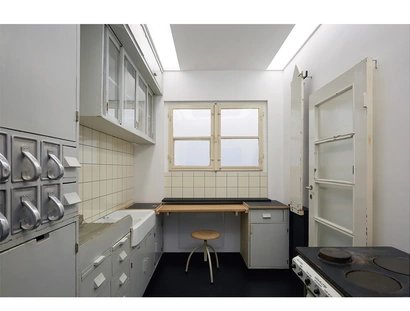 Raumfoto einer Küche mit Herd, Glastür, zugeklapptem Bügeltisch auf der rechten Seite; Schütten für unverpackte Waren aus Metall, Waschbecken und Lagerflächen auf der linken Seite. Im Hintergrund ist eine Arbeitsfläche mit Hocker, Schubladen und Fens