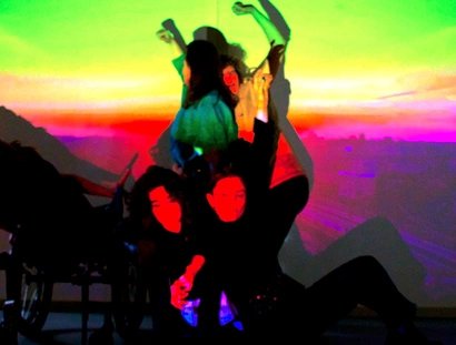 Eine Gruppe von fünf Performer:innen posiert in einer dynamischen, vielschichtigen Komposition. Sie sind vor einer Projektion, die einen leuchtenden Stadthimmel im Sonnenuntergang mit einem Farbverlauf zeigt. Die Projektion wirft starke, gesättigte F