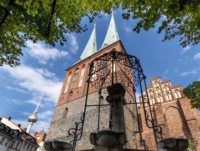 Blick durch eine schmiedeeiserne Brunnenzier auf die Türme und Giebel der Nikolaikirche, im Hinterfgrund hinter den Dächern historisierender Gebäude der Fernsehturm und der Turm des Roten Rathauses