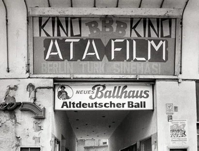 Junge auf Tretroller vor Toreinfahrt in einen Hinterhof, darüber deutsch- und türkischsprachige Werbung