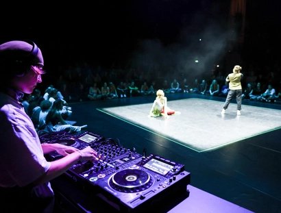 Menschen tanzen auf einer beleuchteten Bühne in einem sonst dunklen im Raum. Im Vordergrund ist angeschnitten ein*e DJ an Turntables zu sehen.
