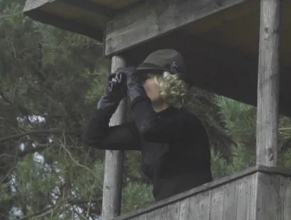 Ein blonde Frau beobachtet auf einem Hochsitz im Wald etwas mit einem Fernglas.
