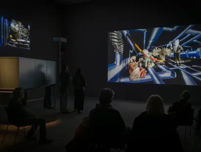 Foto: Ein paar Menschen sitzen und stehen in einem Videoraum und schauen auf eine große Leinwand.