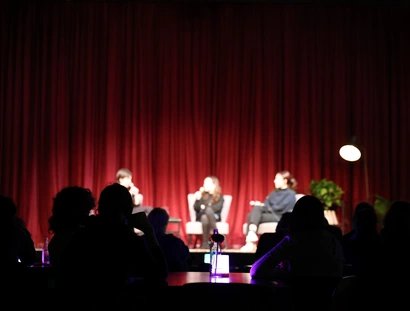 Hinter den Schatten des Publikums sieht man verschwommen drei Personen auf der Bühne vor einem roten Vorhang. Sie sitzen auf Stühlen und halten Mikrofone.