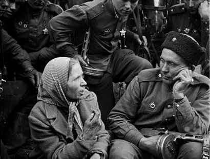 Mehrere Soldaten in Uniform sitzen und stehen neben einem Pferd, eine ältere Frau mit Kopftuch sitzt darunter