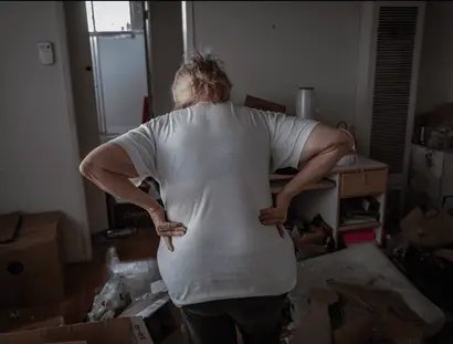 Das Foto zeigt eine Frau, die mit dem Rücken zur Kamera in einem unordentlichen, chaotischen Raum steht. Sie trägt ein weißes T-Shirt und dunkle Hosen und stützt ihre Hände in den unteren Rücken, als sei sie erschöpft oder habe Schmerzen.