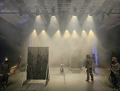 Tänzer:innen in schwarzer Kleidung stehen auf einer nebligen Bühne. Einige blicken nach vorne, andere sind von hinten zu sehen. Zudem befinden sich zwei schwarze Wandaufsteller auf der Bühne.