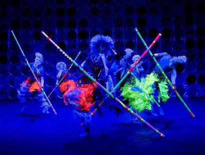 Die rüschigen Kostüme und langen Stäbe von den Tänzer:innen leuchten in bunten Neonfarben.