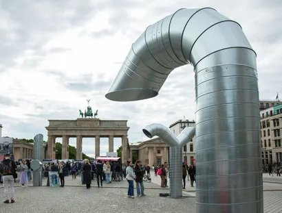 Zwei große, gebogene Metallstrukturen stehen auf dem Pariser Platz vor dem Brandenburger Tor. Menschen versammeln sich in der Nähe.