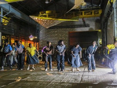 Eine Gruppe von Menschen tanzt energisch in einem großen Veranstaltungsort im Industriestil, der von bunten Lichtern und Discokugeln beleuchtet wird, mit Zuschauern im Hintergrund.