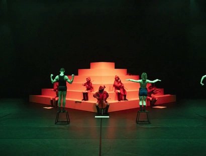 Performer:innen auf Hockern und Musiker:innen in roten Kapuzen posieren vor einer stufenförmigen weißen Plattform im grünen und roten Licht.