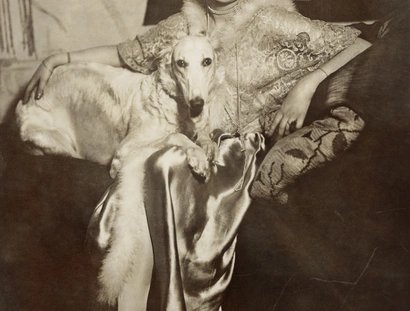 Selbstportrait der Künstlerin Marta-Astfalck-Vietz als Sepia-Fotografie. Sie sitzt in einem glänzenden Kleid mit Pelzbesatz auf einem Sofa. Auf ihrem Schoß sitzt ein Hund mit hellem Fell. Im Hintergrund sind Gemälde und dekorative Elemente zu sehen.