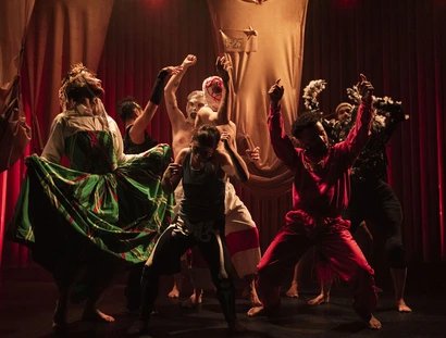 Eine Gruppe von Darstellern in farbenfrohen, dramatischen Kostümen posiert dynamisch auf einer schwach beleuchteten Bühne mit roten Vorhängen und drückt unter der Theaterbeleuchtung intensive Emotionen und Bewegungen aus.