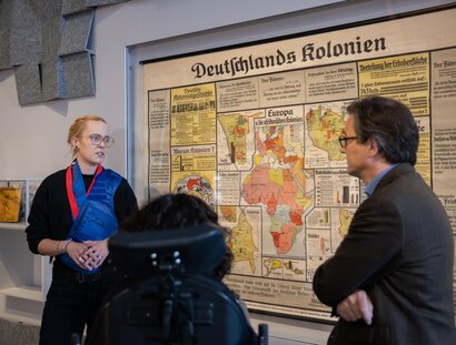 Eine Connectorin erklärt die Hintergründe einer Schulwandkarte aus der Zeit des Nationalsozialismus im Raum "Krieg" der BERLIN GLOBAL Ausstellung