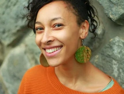 Portrait-Foto der lächelnden Jessica Cuna in organgefarbenem Pullover und mit großen, runden, grün-orangen Ohrhängern