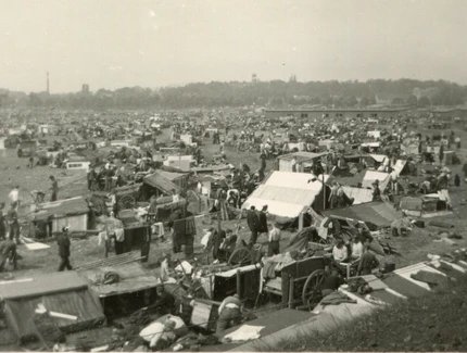 Das Schwarz-Weiß-Foto zeigt eine große Ansammlung von Zelten und Wagen auf einer weitläufigen Fläche. Viele Menschen sind zu sehen, die sich in und um die Behausungen herum bewegen. Im Hintergrund ist eine Bebauung mit Bäumen und Gebäuden erkennbar. 