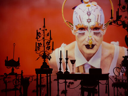 Auf einer rot ausgeleuchteten Bühne befinden sich Kerzenleuchter und ein Tisch, auf eine große Leinwand wird der Performer Steven Cohen in einem fantasievollen Kostüm projiziert.