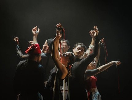 Acht Performer:innen stehen eng beisammen, die Arme anmutig erhoben, rote und grüne Bänder in den Händen – ein kraftvoller, ritueller Moment in dramatischem Bühnenlicht.