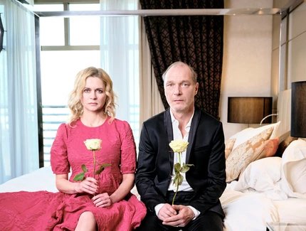 Martin Brambach uind Christine Sommer sitzen auf einem Bett. Beide halten eine gelbe Rose in ihren Händen