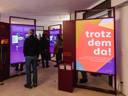 Mehrere Personen betrachten Ausstellungstafeln in einem Raum, eine Tafel zeigt den Schriftzug 'trotzdem da!' in Weiß auf rotem und gelbem Hintergrund.