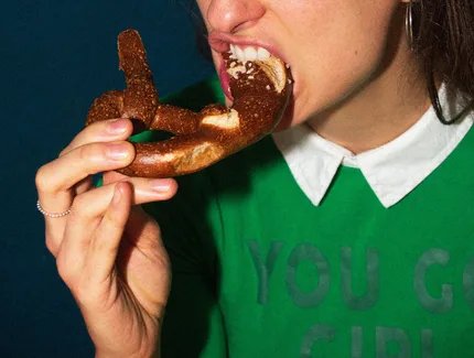 Das Bild zeigt eine junge Frau, die in eine Brezel beißt. Das obere Bildende befindet sich in der Mitte der Nase der abgebildeten Person, sodass ein Großteil des Gesichts und des Kopfes nicht sichtbar sind. Die Person trägt einen grünen Pullover, auf