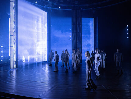 Ein Schauspielensemble steht in gespannter Haltung auf einer blau ausgeleuchteten Bühne.