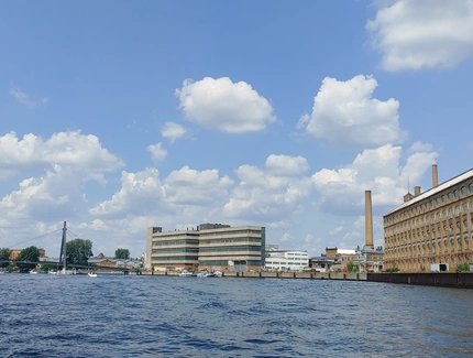 Blick auf einen Fluss mit Industriegebäuden und einem Schornstein unter blauem Himmel mit Wolken